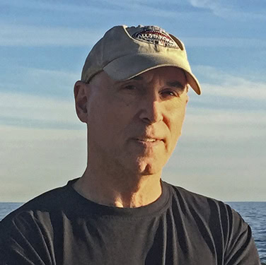 Headshot of Peter wearing a hat with the ocean horizon behind him.