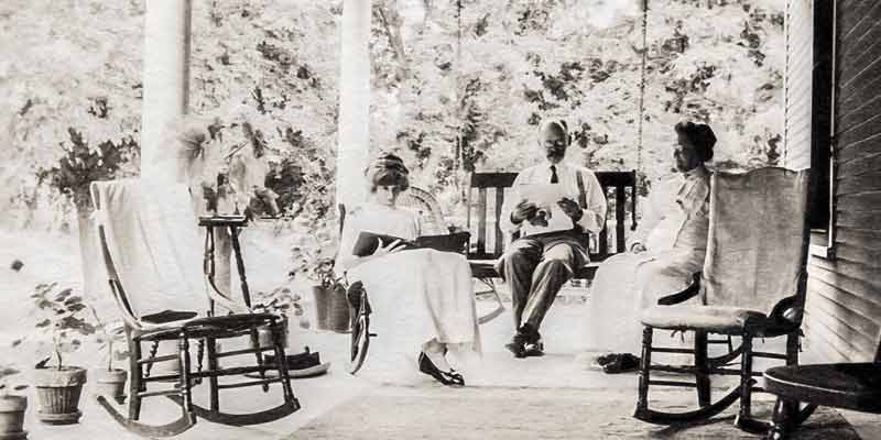 A vintage black and white photo of the Remmers Family sitting on the covered porch reading.